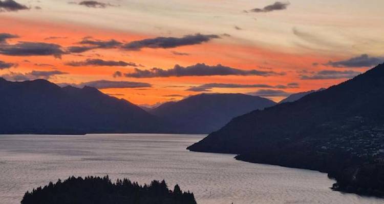 Un coucher de soleil flamboyant peint le ciel au-dessus d'un lac expansif et des silhouettes sombres des montagnes.