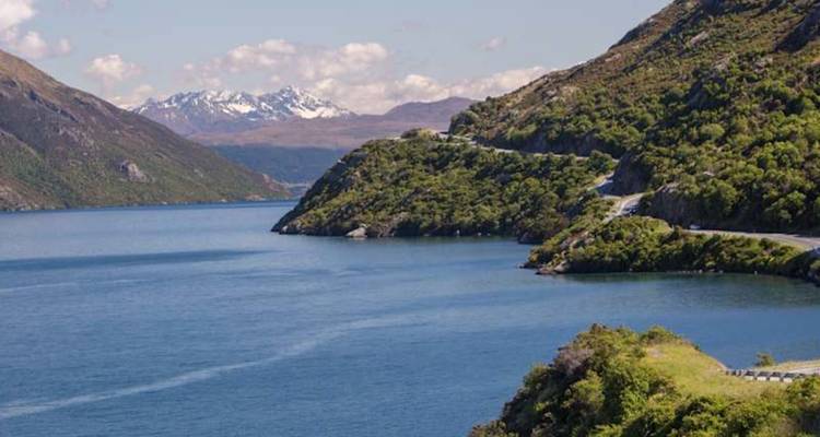 Route de montagne sinueuse longeant un lac bleu profond vers des pics enneigés lointains.