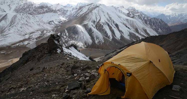 Bright yellow tent perched on a snowy ridge overlooks dramatic jagged peaks and glaciers under moody skies.