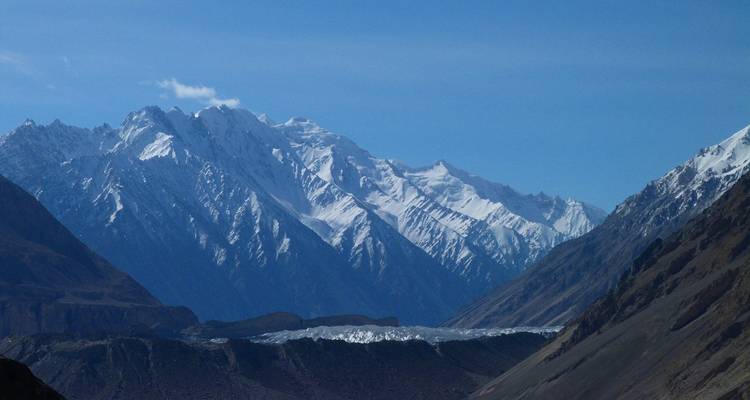 Sharp snow-covered peaks rise above a shimmering glacier under a clear blue mountain sky.