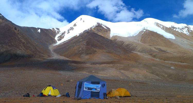 Cluster of bright expedition tents dot a high-altitude plateau beneath snow-rimmed peaks and blue sky.