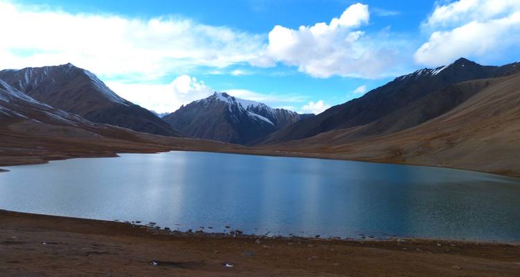 Serene alpine lake mirrors rugged brown slopes and distant snowy summits under scattered clouds.