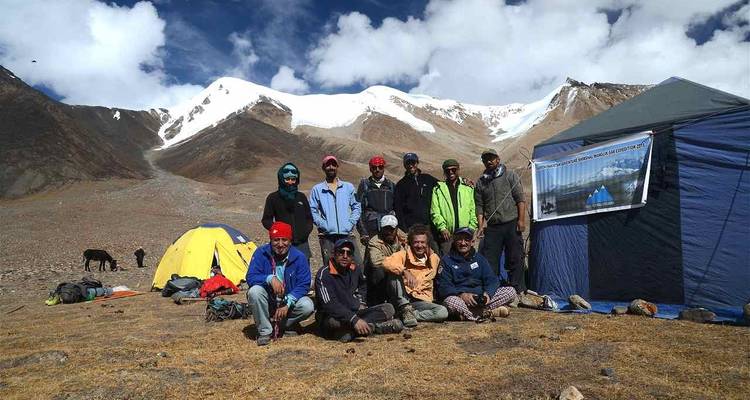 Trekking group poses beside pitched tents at a high mountain base camp against snowy ridges.
