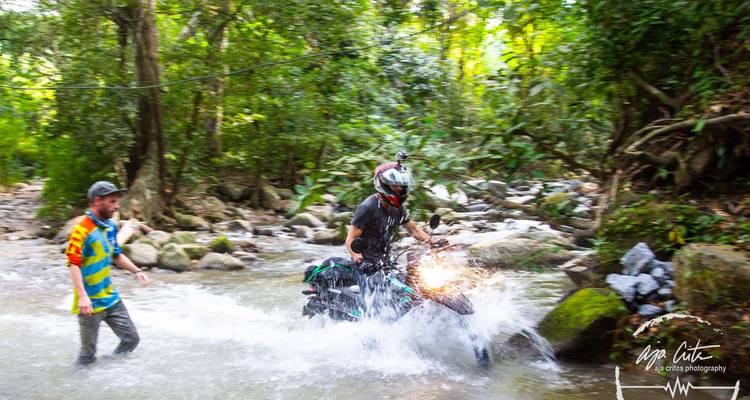 Un motocycliste traverse un ruisseau forestier en éclaboussant l'eau tandis qu'un ami le guide ; filigrane visible.
