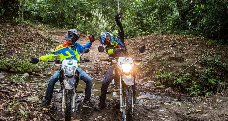 Deux motocyclistes sur des motos tout-terrain font un geste de célébration sur une piste de jungle boueuse à côté d'un petit ruisseau.