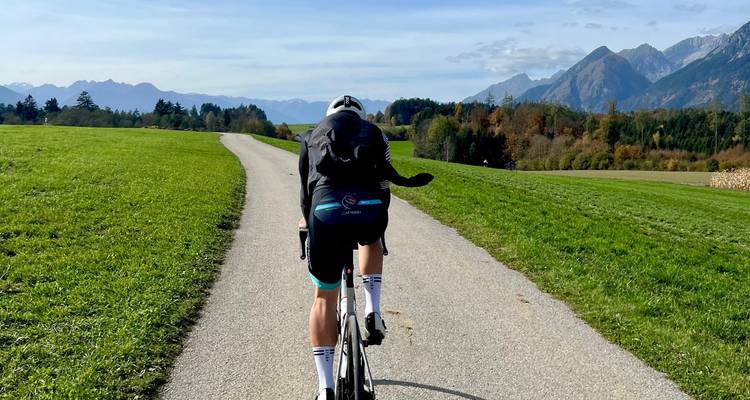 Un cycliste descend rapidement une route pavée rurale à travers des prairies alpines verdoyantes en direction de lointains pics autrichiens.