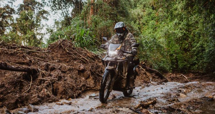 Camouflaged rider navigating a rocky, muddy jungle track on an adventure motorbike.