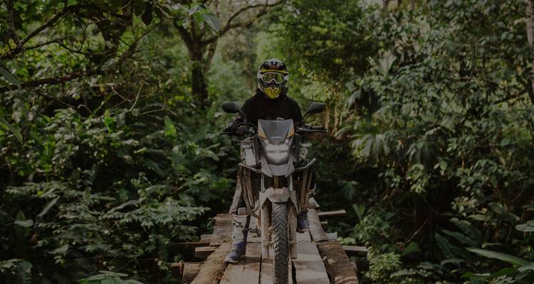Motorradfahrer in vollständiger Schutzausrüstung balanciert auf einer schmalen Holzbrücke tief im Regenwald.