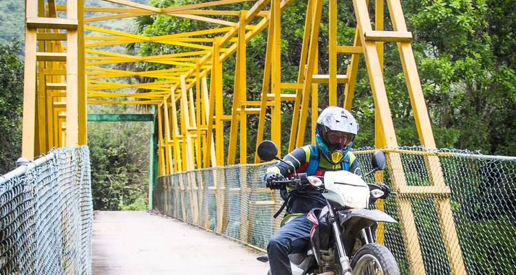 Ein Geländemotorradfahrer überquert eine leuchtend gelbe Fachwerkbrücke in üppiger Dschungelumgebung.