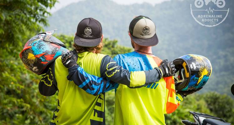 Zwei Motocross-Fahrer in leuchtender Ausrüstung teilen sich eine siegreiche Umarmung mit Bergen im Hintergrund.