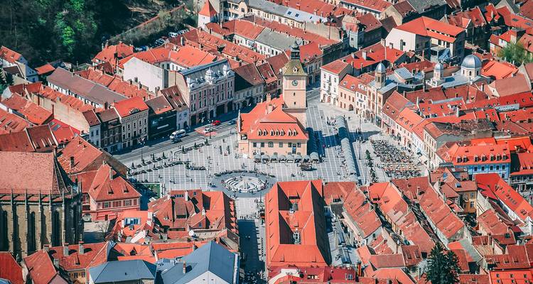 Vue aérienne élevée de la Place du Conseil de Brasov avec des bâtiments médiévaux aux toits rouges nichés dans des collines boisées.