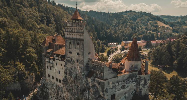 Vue aérienne spectaculaire d'un château médiéval perché sur une colline aux toits rouges entouré d'une forêt dense et verdoyante.