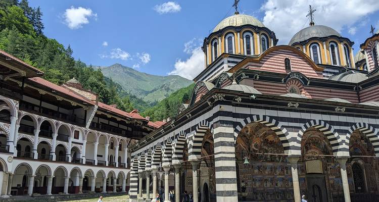 Cloître monastique aux arcades rayées avec des dômes ornés se dressant face aux montagnes boisées et au ciel bleu.