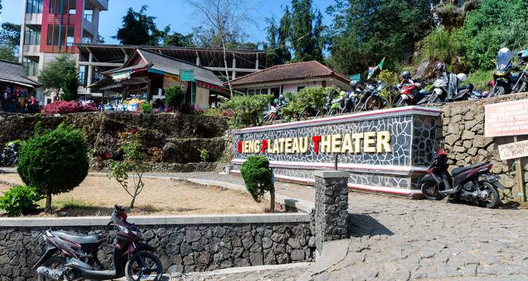Panneau d'entrée du théâtre du plateau de Dieng avec des motos garées et des bâtiments sur la colline