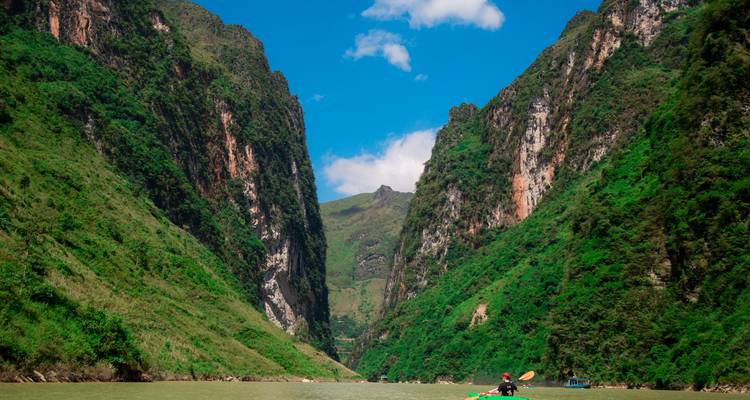 Un kayakista navega con su remo a través de un estrecho cañón de río flanqueado por acantilados verdes imponentes bajo un cielo azul.
