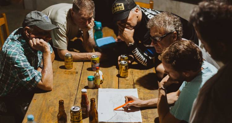 Un grupo de motociclistas se reúne alrededor de un mapa de papel y cervezas mientras planean una ruta de noche.