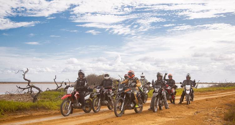 Siete motociclistas se alinean en una pista arenosa bajo un cielo enorme listos para la aventura del desierto.