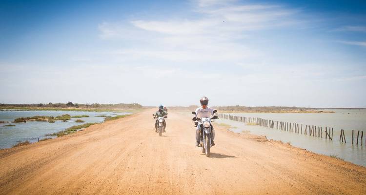 Dos jinetes avanzan rápidamente por una calzada polvorienta flanqueada por lagunas poco profundas.