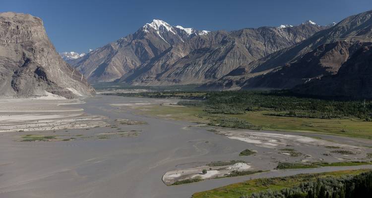 Vaste vallée avec des canaux fluviaux tressés, des prairies verdoyantes et des montagnes de style himalayenne escarpées sous un ciel dégagé.