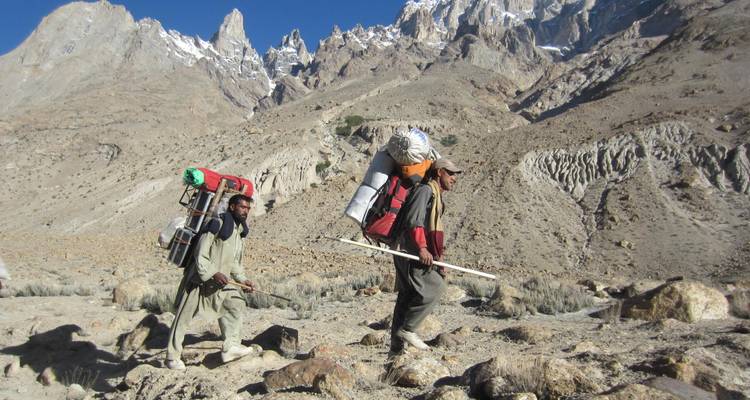 Deux porteurs transportant des charges lourdes le long d'un sentier alpin rocheux avec des pics dentelés en arrière-plan.