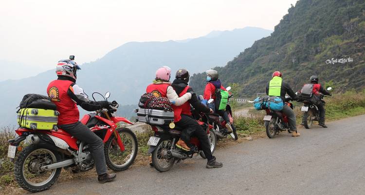 Un grupo de motociclistas con equipo empacado hacen una pausa en un camino de montaña que domina picos brumosos.