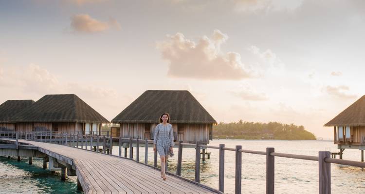 Une femme se promenant le long d'une passerelle en bois menant à des bungalows surplombant l'eau au coucher du soleil.