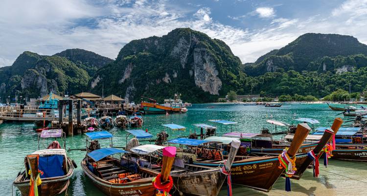 Traditionele longtailboten liggen dicht opeengepakt aan een steiger naast turquoise water en torenhoge jungle-bedekte kliffen op Phi Phi Island.