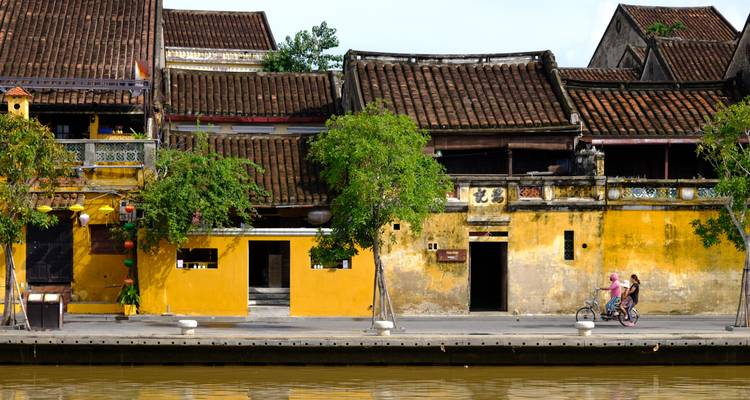Schilderachtige gele winkelhuizen met tegeldaken liggen langs een rivieroever in Hoi An terwijl twee fietsers voorbijrijden.