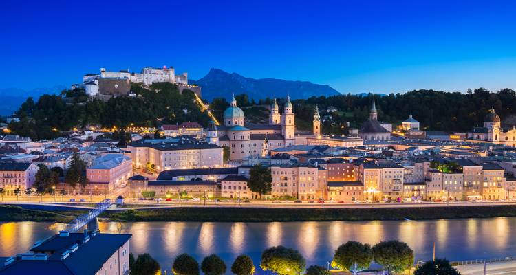 El casco antiguo iluminado de Salzburgo y su fortaleza se reflejan en el río Salzach a la hora azul con los Alpes de fondo.