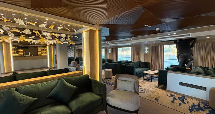 Elegante salón en un barco fluvial con sofás verde oscuro y techo decorativo, un miembro de la tripulación ajustando una ventilación.