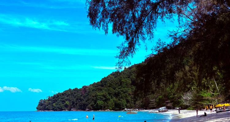 Plage de sable blanc et vaste avec eau turquoise, personnes nageant et pratiquant le paddle-board, en arrière-plan une avancée rocheuse couverte de forêt.