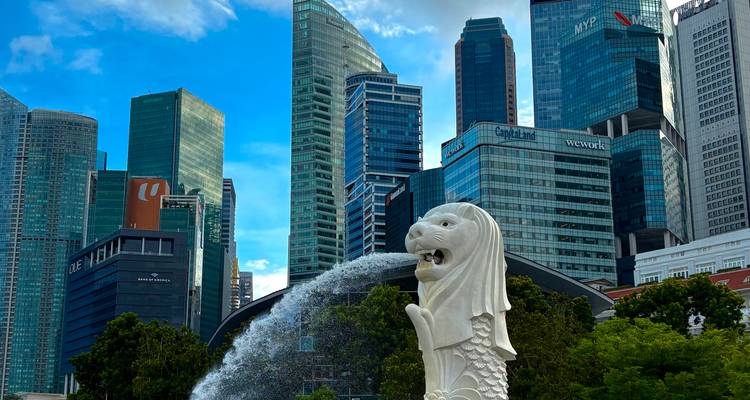 La statue du Merlion à Singapour crache de l'eau en direction de la Marina Bay avec des gratte-ciel modernes en verre qui s'élèvent contre un ciel bleu éclatant.