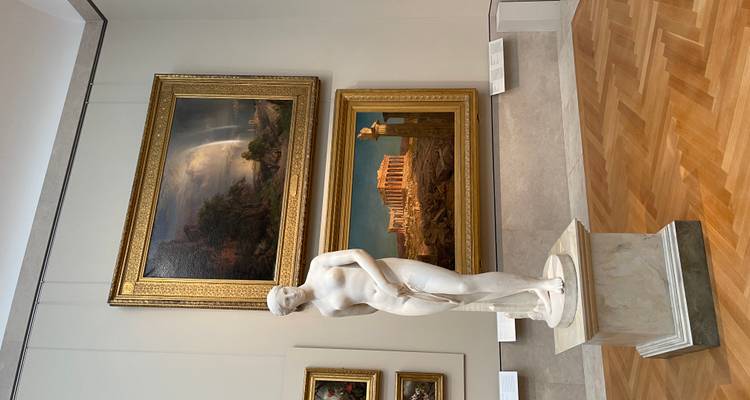 Statue de marbre nu sur un piédestal avec des peintures classiques dans une galerie d'art.