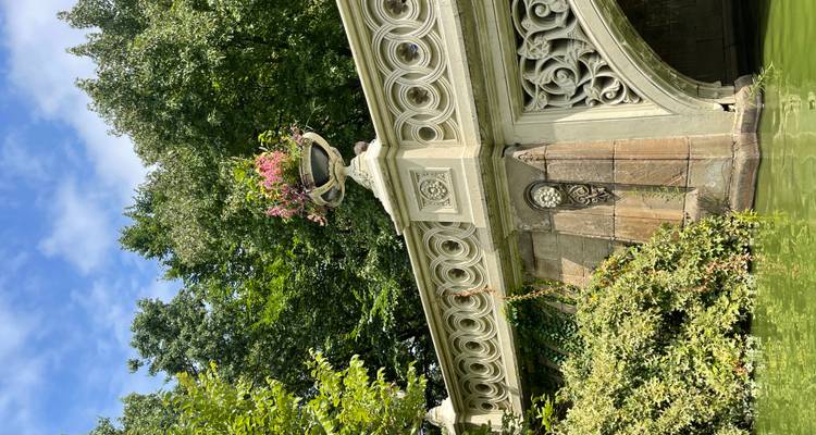 Pont de couleur crème orné avec urne florale enjambant une eau verte entourée d'arbres.