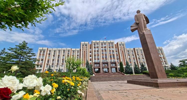 Grand bâtiment gouvernemental de style soviétique avec une statue de Lénine devant, encadré par des parterres de fleurs colorés sous un ciel bleu éclatant.