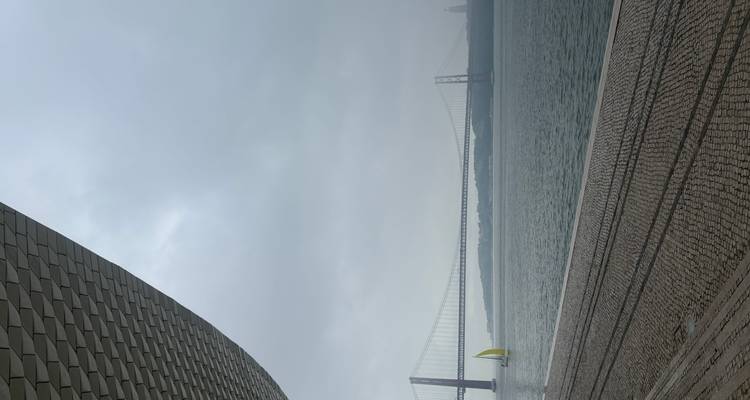 25 de Abril suspension bridge spanning the Tagus with a small yellow sailboat beneath hazy sky