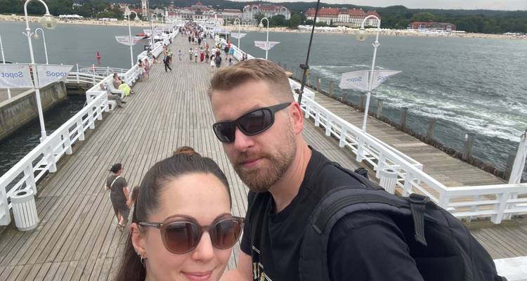 Selfie d'un couple portant des lunettes de soleil sur une longue jetée en bois s'étendant dans la mer avec un complexe hôtelier en arrière-plan.