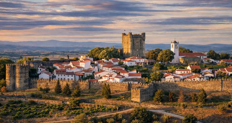 La lumière dorée du coucher de soleil baigne une ville fortifiée sur une colline avec des tours de château médiéval et des toits aux tuiles rouges.