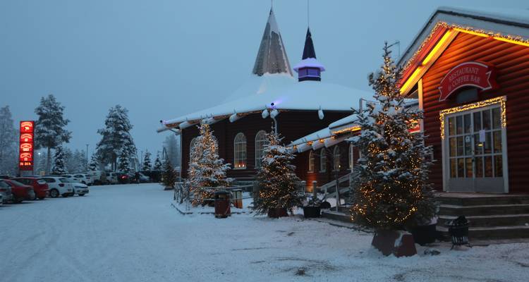 Bâtiments en rondins décorés de lumières de Noël dans un village enneigé au crépuscule.