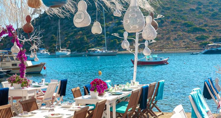 Restaurant en plein air en bord de mer avec des tables blanches, des chaises turquoise et des lanternes décoratives à côté d'une baie calme avec des yachts mouillés.