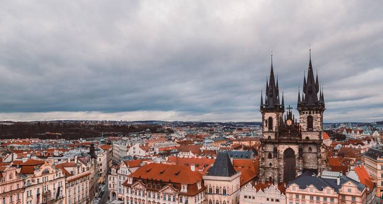 Vista panorámica desde la azotea del Casco Antiguo de Praga con las dos torres de la Iglesia de Týn elevándose sobre las casas de tejado rojo bajo un cielo nublado.