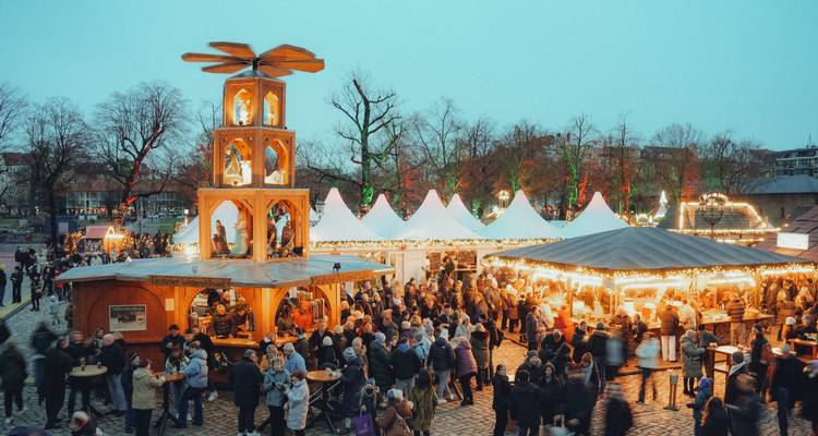 La gente pasea por los puestos brillantes de un animado mercado navideño al aire libre rodeado de carpas blancas y torres de madera.
