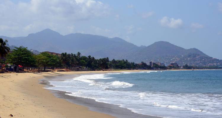 Una playa tranquila y arenosa con olas suaves y montañas que se elevan de fondo cerca de Freetown.