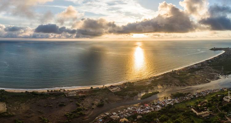 Panorámica con dron de una bahía extensa al atardecer, con la luz dorada del sol reflejándose en el océano y nubes dramáticas en el cielo.