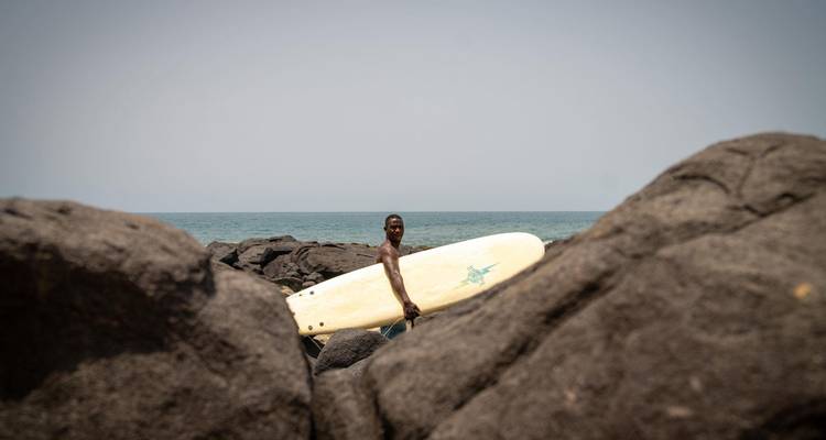Surfista llevando tabla caminando entre rocas volcánicas hacia el océano bajo un cielo brumoso.