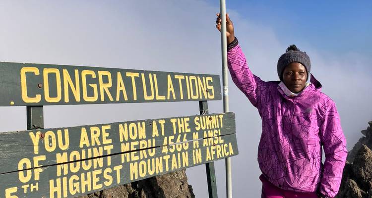 Un alpiniste vêtu d'une veste rose pose à côté d'un panneau au sommet indiquant « Félicitations, vous êtes maintenant au sommet du Mont Meru ».