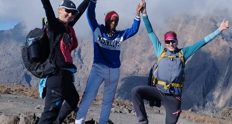 Trois randonneurs joyeux prennent des poses célébratoires sur un versant alpin rocheux avec le rebord du cratère du Mont Meru derrière eux.