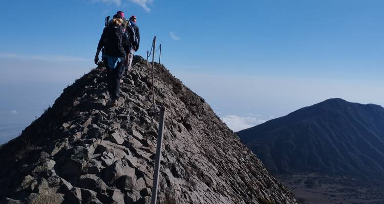 Ligne de randonneurs négociant une crête rocheuse étroite en direction du sommet du Mont Méru avec de vastes vallées en contrebas.