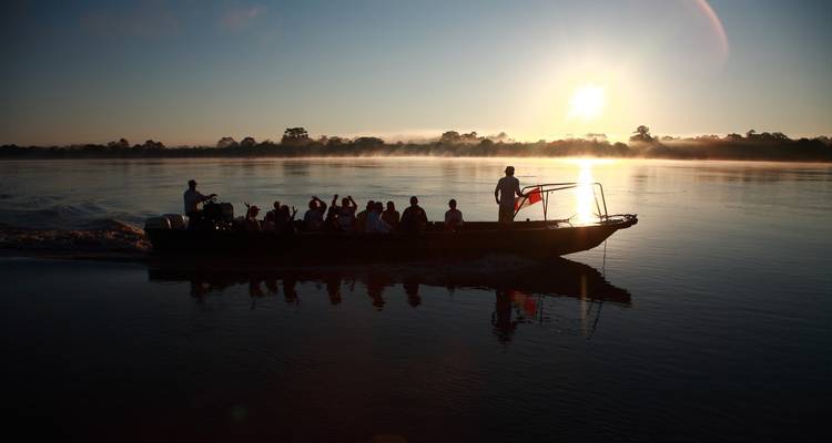 Des passagers en silhouette traversent des eaux fluviales brumeuses à bord d'un bateau rapide au lever du soleil, le soleil brillant bas à l'horizon.