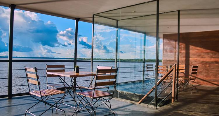 Pont ouvert avec table de bistrot en bois offrant une vue panoramique sur le fleuve et les nuages dramatiques à travers les parois de verre.